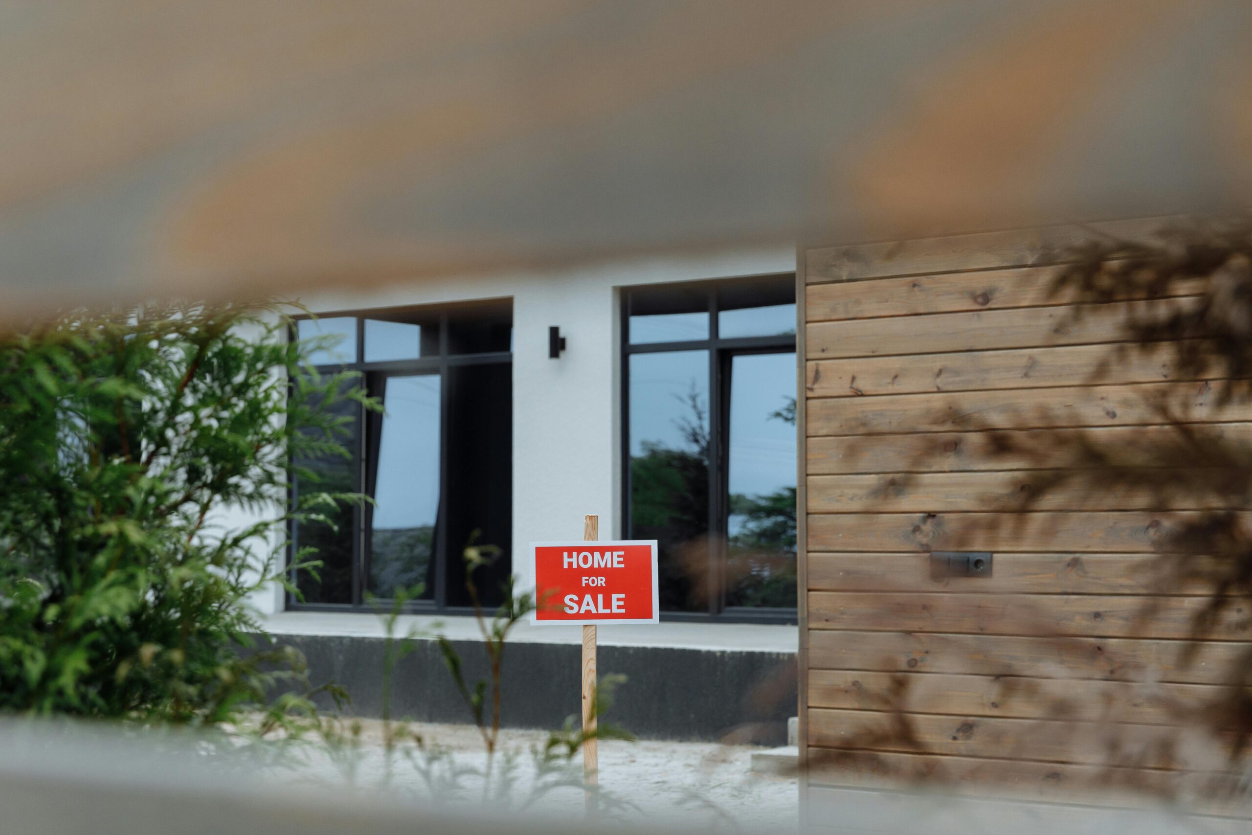 A contemporary home with a 'For Sale' sign in the front yard, framed by greenery.